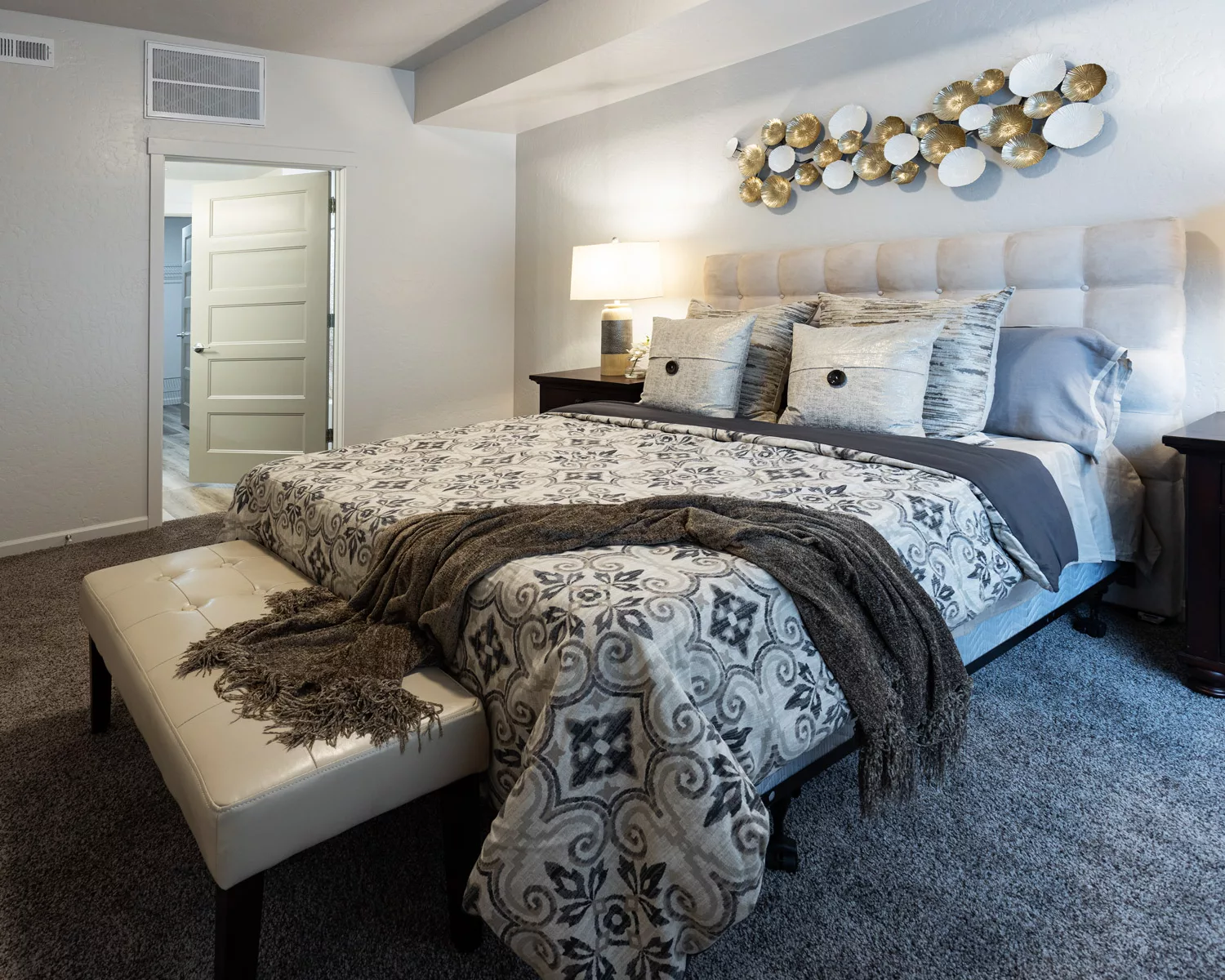 A neatly arranged bedroom featuring a patterned bedspread, plush cushions, a bench at the foot, and decorative wall art.
