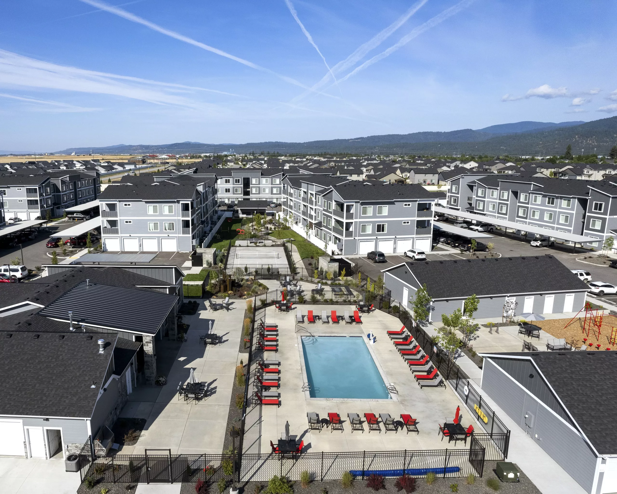 Aerial view of a residential complex featuring several modern, gray buildings surrounding a central pool with red lounge chairs.