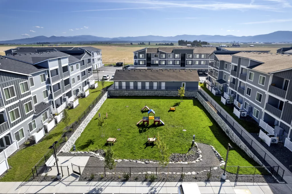 An aerial view of apartment buildings surrounding a grassy courtyard with a small playground and picnic tables, against a backdrop of open fields and mountains.