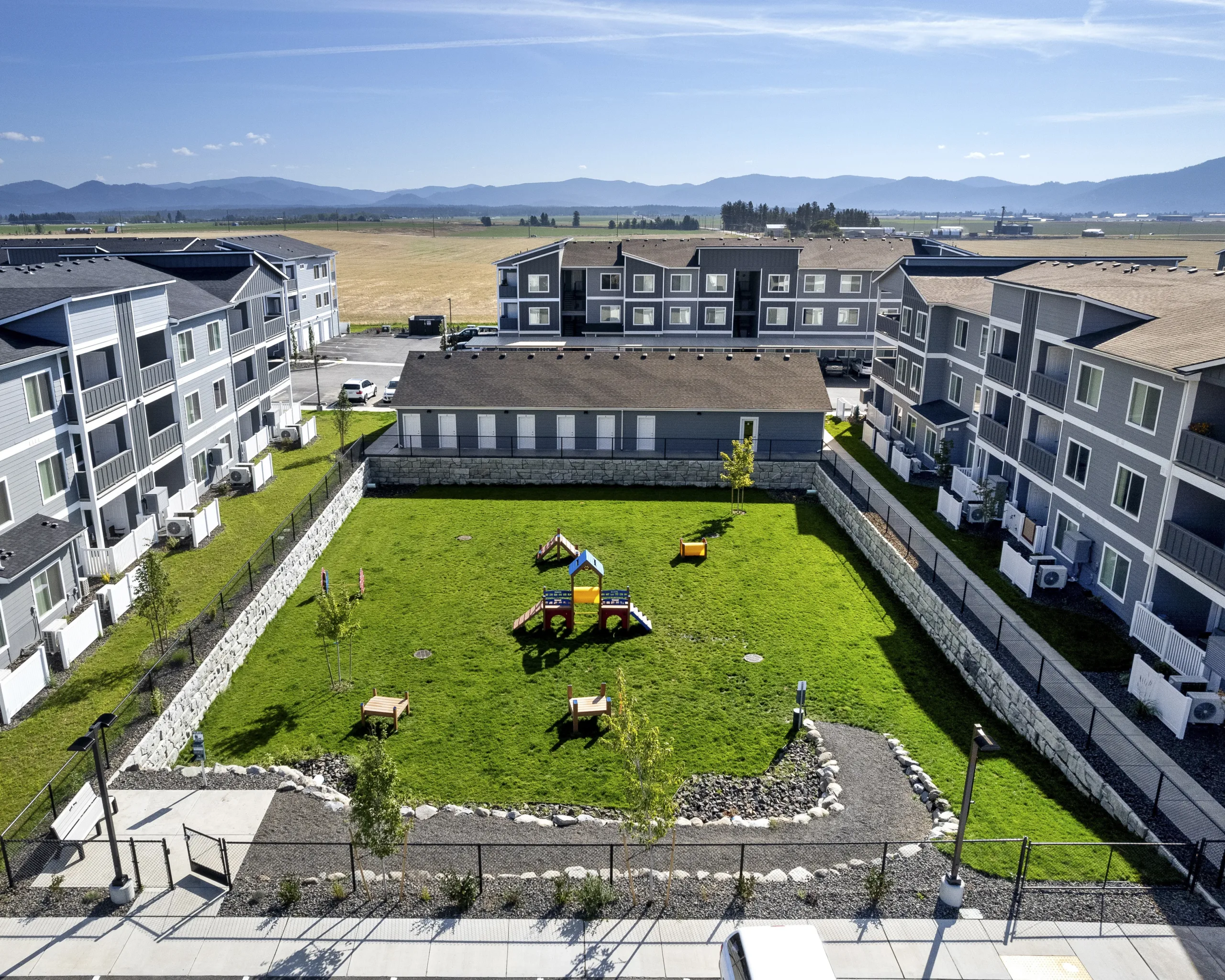 Ariel view of an enclosed green space with playground equipment, surrounded by modern residential buildings and open fields in the background.